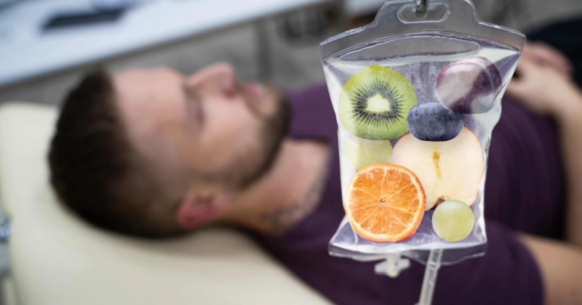 A blurry man in a purple shirt lies on his back, while a clear IV bag filled with fruits (kiwi, orange, grapes, blueberry) hangs in a clinic setting for IV Vitamin Therapy in Buford, GA.